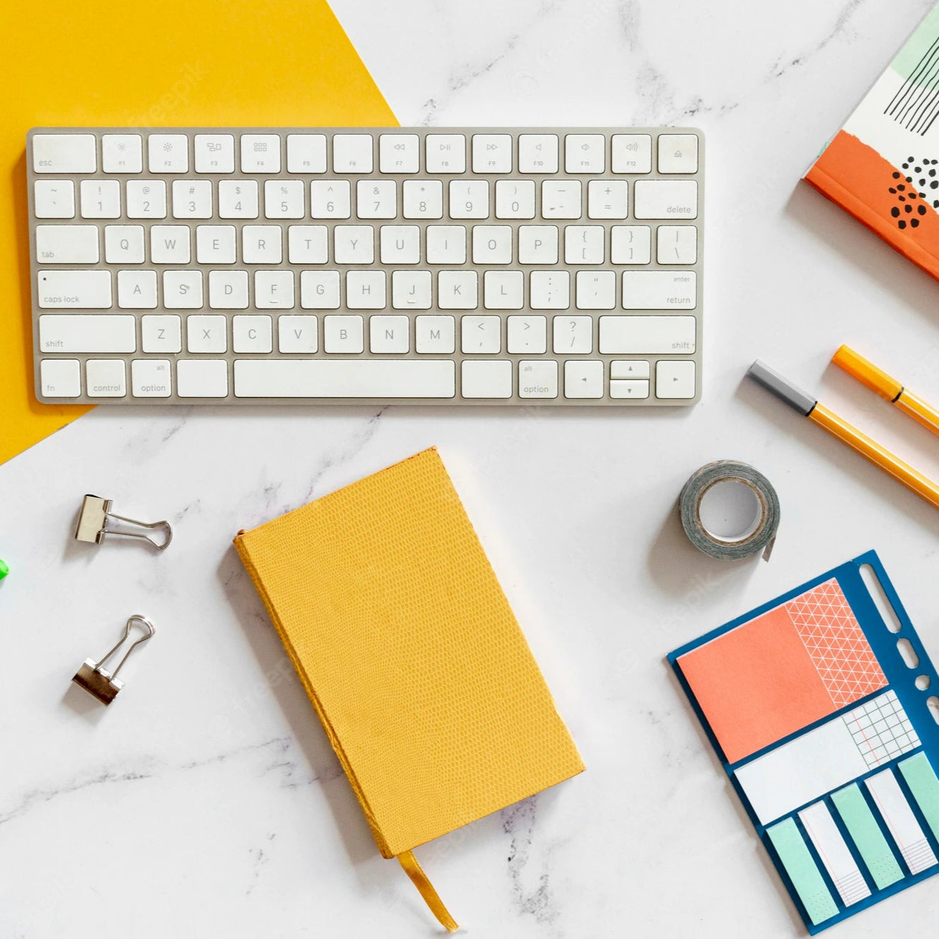 Keyboard, notebooks, pens, and office supplies on a white marble surface with a yellow accent.