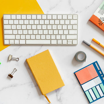 Keyboard, notebooks, pens, and office supplies on a white marble surface with a yellow accent.